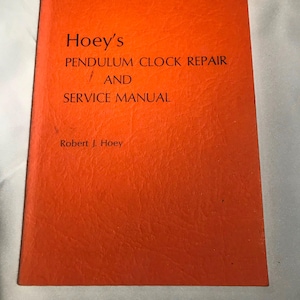 May include: An orange book titled "Hoey's Pendulum Clock Repair and Service Manual" by Robert J. Hoey. The book's cover has a textured appearance. Handwritten text and a price are visible in the upper right corner.