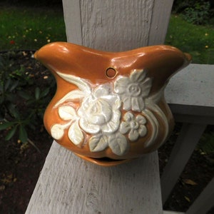 May include: A brown ceramic wall planter with a white floral design. The planter has a hole in the back for hanging.