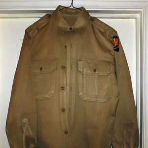 May include: A tan long-sleeved shirt with a button-up front and two chest pockets. The shirt has a patch on the right sleeve with a gold eagle design.