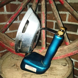 Puede incluir: Una plancha azul y plateada vintage con un mango curvo y un accesorio de latón. La plancha está sobre un barril de madera. Una rueda de carro roja y de madera está en el fondo.