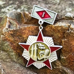 May include: A vintage military medal with a red star and a central emblem featuring a gold design. The medal has a rectangular top piece with a red diamond and the number "I". The medal is set against a textured, brown stone background.