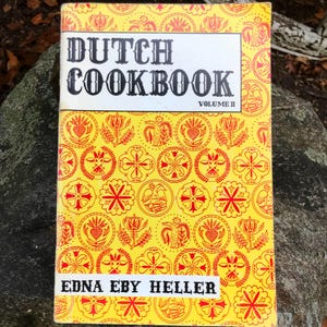 May include: A vintage cookbook titled "Dutch Cookbook Volume II" with a yellow cover featuring red floral and geometric patterns. The title is in bold black letters, and the author's name, Edna Eby Heller, is printed below the title.