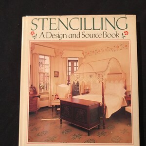 Può includere: Un libro intitolato "Stencilling: A Design and Source Book" con una foto di una camera da letto con un letto a baldacchino, un baule e una sedia. Le pareti e il pavimento sono decorati con stencil.