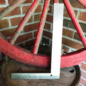 May include: A weathered, red wooden wagon wheel rests on a wooden barrel. A metal ruler is placed on the barrel and against the wheel. The wheel's spokes and rim show signs of age and wear, with visible paint chipping.