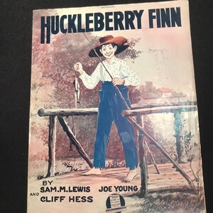 Op de afbeelding: Een vintage bladmuziekhoes met een cartoonillustratie van Huckleberry Finn die aan het vissen is op een houten brug. De titel "Huckleberry Finn" staat in grote, vetgedrukte letters bovenaan de hoes. De muziek is van Sam M. Lewis en Joe Young.