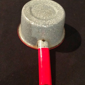 May include: A small, speckled grey and red saucepan with a bright red handle. The pot has a rounded body and a red rim. The handle is tubular with a hole at the end for hanging. The saucepan is likely for cooking or heating small portions.