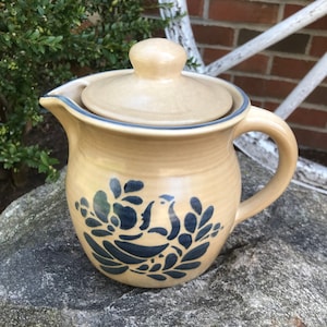 May include: A tan ceramic pitcher with a blue and white floral design. The pitcher has a lid and a handle.