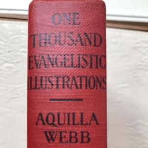 May include: A red book spine with the title "ONE THOUSAND EVANGELISTIC ILLUSTRATIONS" and the author's name, "AQUILLA WEBB," in black lettering. The book has a textured cover.