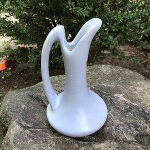May include: A light blue ceramic pitcher with a unique, curved handle. The pitcher has a smooth, matte finish and a wide base.