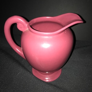 May include: A vintage, deep pink ceramic creamer jug with a rounded body and a curved spout. The jug has a smooth, glossy finish.