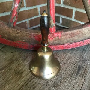 May include: A brass bell with a wooden handle. The bell is polished and has a shiny finish. The handle is dark brown and has a gold-colored top.