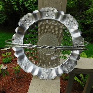 May include: A decorative, vintage-style serving dish with a scalloped, silver-colored rim and a clear glass center. The rim features an embossed design. Two silver handles are attached to the rim, and a twisted metal bar spans the center.