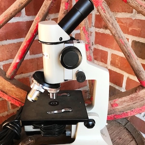 May include: A white and black microscope with a black cord and a light bulb. The microscope is sitting on a wooden barrel. The brand name "BAUSCH & LOMB" is visible on the base of the microscope.