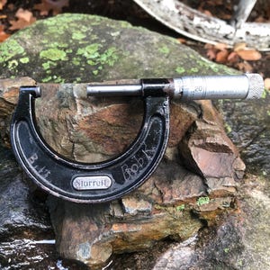 May include: A black Starrett micrometer sits on a rock. The micrometer has a silver measuring scale and the name "Bob K" is etched on the black frame. The rock is brown and grey with patches of green.
