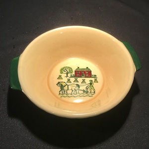 Puede incluir: Un tazón de cerámica de color crema con asas verdes y una escena pintada de una granja roja, un carro tirado por caballos y un granjero. El tazón es perfecto para servir sopa o cereal.