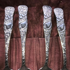 May include: Five silver spoons with ornate, detailed designs. The spoons are arranged in a row, with the handles facing up. The spoons are in a brown leather case.