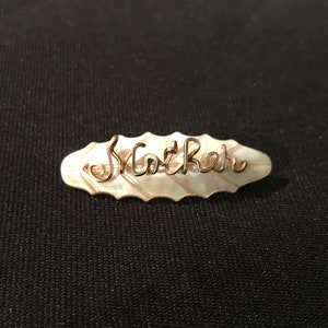 Puede incluir: Un broche ovalado blanco con una inscripción de alambre dorado que dice "Mar Ree".