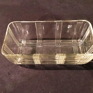 Puede incluir: Un plato rectangular de vidrio transparente con un patrón de cuadrícula grabado en la base y los lados. El plato tiene un borde ligeramente elevado y está diseñado para servir o exhibir. El vidrio es transparente.