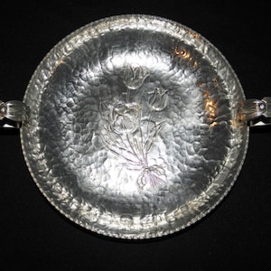 May include: A silver hammered metal serving tray with two handles and a floral design. The tray has a textured surface and a beaded edge.