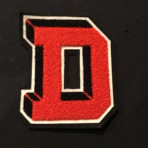 May include: A red letter "D" patch with a white border and black shadow effect. The letter has a textured, felt-like appearance. The patch is likely for a jacket or other apparel.