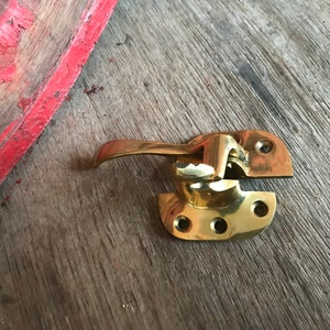 May include: A brass window latch with a lever handle. The latch is in two pieces, with the handle attached to the top piece. The latch is on a wooden surface.