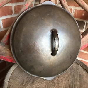 May include: A vintage cast iron pot lid with a single handle. The lid is dark gray and has a smooth, slightly textured surface.