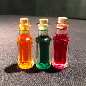 Könnte beinhalten: Drei kleine Glasflaschen mit Korkstopfen. Die Flaschen enthalten orange, grüne und rote Flüssigkeiten.