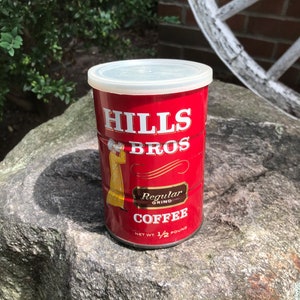 Könnte beinhalten: Rote und weiße Hills Bros. Kaffeedose mit weißem Deckel. Die Dose zeigt ein Bild einer Person in einem gelben Mantel und den Text "Regular Grind Coffee" und "Net Wt. 1/2 Pound".