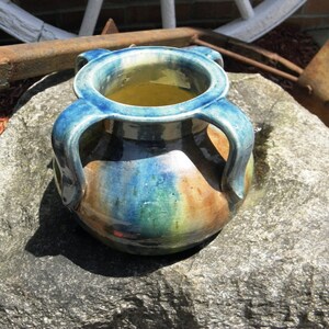 May include: A handmade ceramic vase with two handles, featuring a blue rim and handles, with a body that transitions from brown to green and blue. The vase is sitting on a gray rock.
