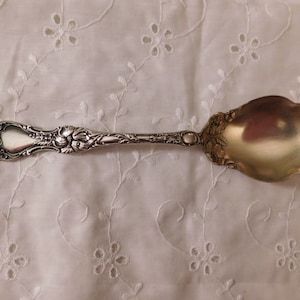 Puede incluir: Una cuchara de azúcar de plata con un mango adornado y un cuenco redondeado. La cuchara tiene un diseño floral y está en buen estado.