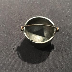 Może przedstawiać: Małe, srebrne wiaderko z metalu z drucianym uchwytem. Wiaderko ma cylindryczny kształt i jest lekko przyciemnione, co nadaje mu wygląd vintage. Uchwyt jest przymocowany za pomocą małych, okrągłych łączników.