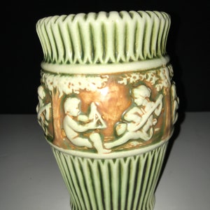 May include: A green and white ceramic vase with a brown band featuring a relief design of two figures playing a game. The vase has a fluted design around the top and bottom.