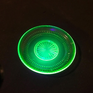 May include: A round, glowing green glass dish with a central starburst design. The dish has a series of concentric circles and a textured rim. The glass appears to be vintage, with a bright, almost neon, green color.