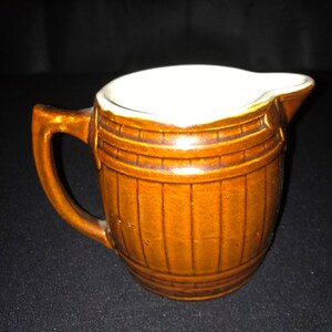 May include: A brown ceramic creamer shaped like a barrel with a white interior and a handle.