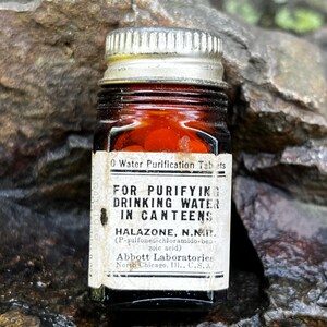 Könnte beinhalten: Eine kleine bernsteinfarbene Glasflasche mit silbernem Metallschraubverschluss, die rote Tabletten enthält. Das weiße Papieretikett trägt die Aufschrift: Water Purification Tablets, FOR PURIFYING DRINKING WATER IN CANTEENS, HALAZONE, N.F., Abbott Laboratories.