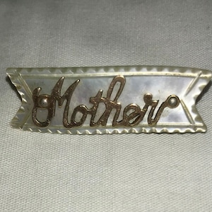 May include: A white mother's day pin with gold lettering that reads "Mother".