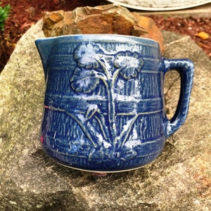 Puede incluir: Una jarra de cerámica azul con un diseño floral. La jarra tiene un asa y una boca ancha.