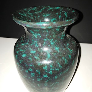 May include: A dark green vase with a speckled pattern of teal and dark blue. The vase has a wide, flared opening and a rounded body. The surface is glossy, reflecting light. The vase is a decorative item.