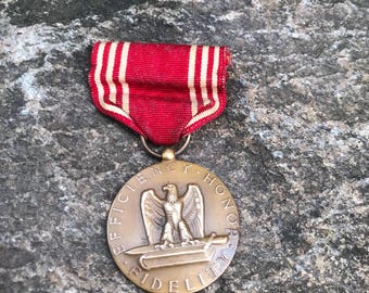 Könnte beinhalten: Eine Bronzemedaille mit einem rot-weiß-roten Band. Die Medaille zeigt einen Adler, der auf einem Buch mit einem Schwert sitzt. Die Inschrift auf der Medaille lautet "Effizienz, Ehre, Treue".