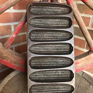 Puede incluir: Molde de pan de maíz de hierro fundido con forma de seis mazorcas de maíz. La sartén gris oscuro tiene un asa. El fondo incluye una rueda de carro roja y una pared de ladrillos.