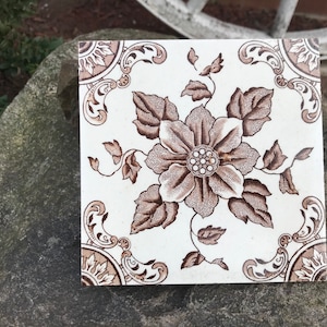 May include: A vintage ceramic tile with a brown and white floral design. The tile features a large flower with intricate details and leaves.