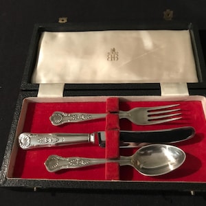 May include: A black box with a red velvet interior containing a silver fork, knife, and spoon. The box has a label with the text "Royal Crown" and the initials "CHB".
