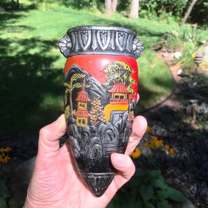 Pode incluir: Um vaso de cerâmica preto e vermelho com um design asiático detalhado. O vaso apresenta uma cena de montanha com pagodas, árvores e um céu vermelho e amarelo. O vaso tem uma superfície texturizada e uma borda prateada.