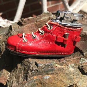 Puede incluir: Un encendedor novedoso con forma de zapato de cuero rojo con cordones blancos. El encendedor con forma de zapato tiene una parte superior de metal plateado y está colocado sobre una roca rugosa y texturizada. El encendedor es un artículo de colección único.