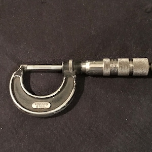 Puede incluir: Un micrómetro plateado y negro con un mango negro y un mecanismo de medición plateado. El mango tiene las palabras "FORGED STEEL" impresas.