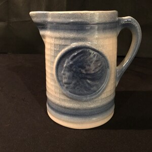 May include: Blue and white ceramic pitcher with a Native American profile in a circle on the front. The pitcher has a handle and a rounded base.