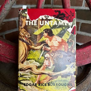 Peut inclure: Un livre relié vintage intitulé Tarzan the Untamed par Edgar Rice Burroughs. La jaquette présente une illustration colorée d'un homme tenant un couteau, un léopard et un lion dans un décor de jungle. La couverture montre des signes d'usure.