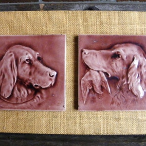 May include: Two brown ceramic tiles with raised relief images of dogs. The tiles are framed in a brown burlap mat.