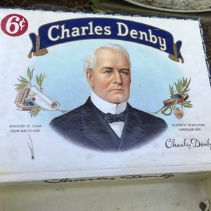 Könnte beinhalten: Eine Vintage-Zigarre-Box mit einem Porträt von Charles Denby, einem ehemaligen US-amerikanischen Minister in China. Die Box ist weiß mit blauen und goldenen Akzenten. Der Text auf der Box lautet "6¢ Charles Denby", "Minister für China von 1885 bis 1898", "Mitglied der Philippinischen Kommission 1899" und "Charles Denby".