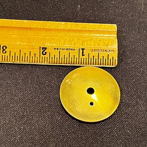 Op de afbeelding: Een ronde, goudkleurige metalen knoop met een glad, licht concaaf oppervlak en twee kleine gaatjes voor bevestiging. De knoop ligt onder een gele houten liniaal die een diameter van ongeveer 3,2 cm aangeeft.